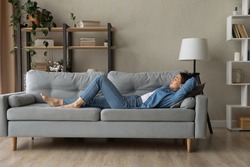 Side view woman enjoy day nap on comfy sofa. Young caucasian female put hands behind hear lying on cushion on cozy couch breath fresh conditioned air inside modern living room. Leisure, repose concept