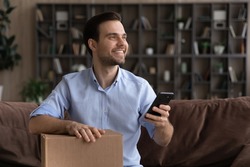 Satisfied millennial man sit on couch hold postal parcel smartphone enjoy express delivery courier service ordered by phone call. Happy young male client give positive feedback to package tracking app