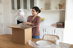 Happy young Indian female renter or tenant settle in new home unbox parcel shopping tableware online from home. Smiling ethnic woman unpack box buying kitchenware on internet. Delivery concept.