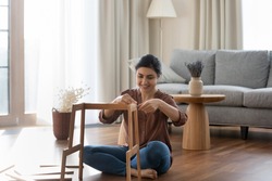 Happy millennial Indian female renter or tenant sit on floor in new house gather shelf. Smiling young ethnic woman put together make build piece of furniture settle in own home. Rental concept.