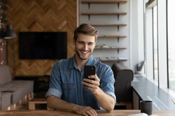 Happy joyful millennial handsome guy taking picture on smartphone, using online app on gadget. Young man making video call from home, talking via virtual conference chat, smiling at cellphone camera
