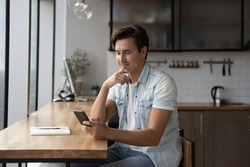 Happy thoughtful millennial guy using app, online service on smartphone at home workplace table, looking at screen, browsing internet, reading text, typing message. Man making call on mobile phone