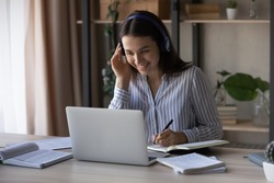 Smiling millennial Caucasian woman in headphones sit on table at home study distant on computer. Happy young female in earphones have webcam online video class or lesson on laptop. Education concept.