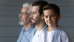 Portrait of happy small teen boy look at camera pose on grey wall background with young Caucasian father and old grandfather. Three generations of men show family unity and boding. Offspring concept.