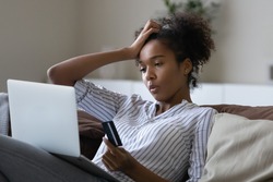 Unhappy millennial African American woman distressed with problems paying online on laptop with credit debit card. Upset young ethnic female confused have troubles shopping on web on computer.
