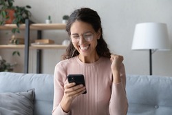 Attractive happy Latina woman in glasses sit on sofa at home holding smartphone read sms notification great news looking excited overjoyed cant believe in luck enjoy moment of victory. Success concept