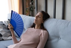 Overheated tired young Latina woman sit lean on sofa at home wave hand fan reduce heat, suffer from hot weather, lack of air conditioner indoor feels unwell exhausted, have hormonal imbalance concept