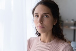 Close up portrait of joyless 30s woman standing alone near window indoor. Face of Hispanic serious female with sad eyes staring at camera. Tiredness, lack of optimism, solitude, life concerns concept