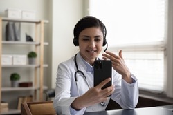 Happy young indian general practitioner therapist in headset holding video call web camera conversation on cellphone, communicating distantly with patient or e-learning on virtual conference event.