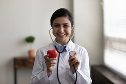 Smiling young professional female indian ethnicity cardiologist wearing stethoscope, showing heart figure to camera, reminding regular examination checkup meeting, cardiovascular disease prevention.