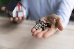 Hand of realtor giving key from new home, holding tiny toy model of house, offering apartment for rent, buying. Homeowner, tenant buying, renting property. Real estate, mortgage concept. Close up