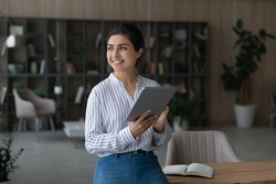 Dreamy smiling Indian businesswoman holding tablet, looking to aside, standing in home office, happy young female visualizing future, dreaming about new job opportunities, distracted from work