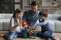 Happy best dad and two sibling children playing together on warm floor, building toy tower on clean carpet, constructing model from plastic blocks, improving creativity in game. Family home activity