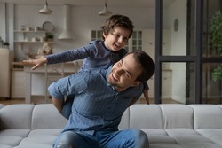 Happy loving daddy and affectionate boy playing active games at home. Dad piggybacking son on couch, kid making airplane wings with open flying hands, laughing, having fun. Family leisure concept