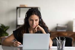 Focused female adult student in headphones using laptop, attending online lesson, virtual class, listening webinar, watching training, writing notes, making video call. Remote learning on internet
