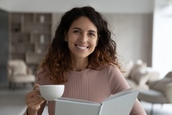 Portrait of smiling young latin woman holding mug of hot coffee or tea, reading favorite literature novel paper book, enjoying peaceful carefree leisure hobby activity pastime on weekend alone at home