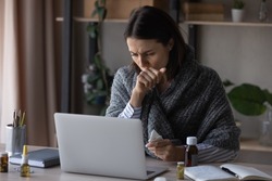 Unhealthy businesswoman wrapped in plaid working on laptop at home office, feeling unwell, sick young woman freelancer or student sitting at desk with medications, suffering from flu or virus