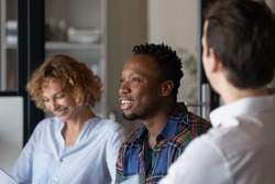 Diverse team of office employees discussing project together, brainstorming, talking, laughing. Smiling African American guy visiting group therapy meeting, sharing funny story to laughing groupmates