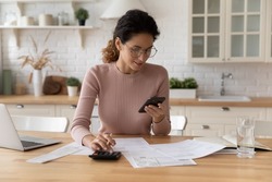 Happy young Caucasian woman sit at desk at home manage household expenses expenditures paying on cellphone online. Millennial female calculate budget finances, make payment on smartphone.