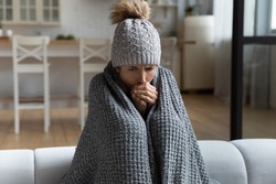 Unwell millennial female renter in hat and blanket sit in cold living room suffer from air conditioner lack. Unhealthy young Caucasian woman struggle from chill freeze at home. No heating concept.