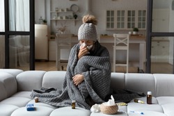 Unhealthy woman sit in blanket hat feel sick suffer from flu or covid-19 corona virus. Unwell young Caucasian female sneeze sniffle have running nose, struggle with influenza or cold at home.