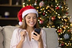 Happy overjoyed Xmas girl in Santa cap excited with good news about gift, looking at smartphone screen, reading text, chatting online, celebrating Christmas holiday, enjoying New Year congratulations