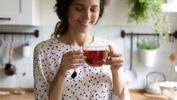 Happy woman taking tea break, holding glass mug, standing in kitchen, smelling and drinking hot aromatic beverage, enjoying morning and breakfast at home. Teatime, hygge, healthy lifestyle concept