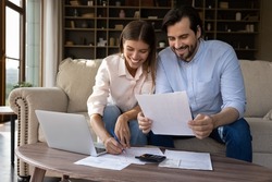 Happy young family couple managing household budget, enjoying planning investment, analyzing paper bills, making payments online in e-banking computer application, doing financial paperwork at home.