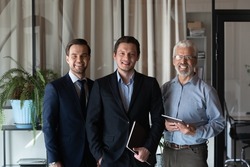 Smiling friendly group of three businessmen of different age private company staff posing for office portrait. Young male team leader ceo and two men aged and millennial subordinates looking at camera