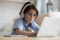 Serious gen Z boy in headphones studying from home, watching online lesson on laptop, attending distance virtual class, making video call, listening to school teacher. Child education concept