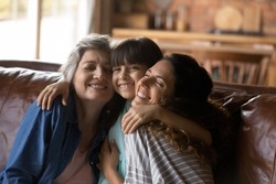 Three female generations family portrait. Cute girl hugging tightly beloved mom and grandma. Mother and grandmother embracing, cuddling granddaughter on couch at home, enjoying leisure time together