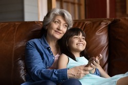 Happy grandmother hugging grand kid on couch at home, holding 7s girl in arms. Grey haired senior grandma enjoying leisure time with laughing cute granddaughter, talking to child. Family relations