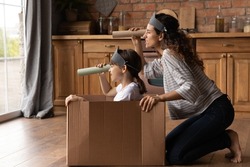 Happy creative mother and active daughter girl playing pirates ship at home, sailing carton box boat, looking forward through toy paper spyglasses, enjoying home activities. Family role game