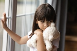 Sad lonely kid holding, hugging teddy bear toy, standing at window alone, feeling depressed, upset, unhappy, going through trauma, touching glass. Childhood problems, abuse, in family concept
