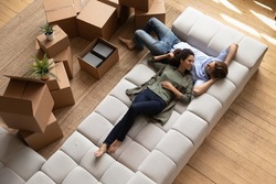 Overhead view peaceful young couple relaxing on fashionable cozy sofa near heap of packed belongings in cardboard boxes, resting at own house at relocation day. Bank loan and mortgage, tenancy concept