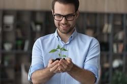 Happy dreamy young handsome ceo executive manager in eyewear holding in hands small green plant, symbol of developing new successful project, eco-friendly sustainable company growth path concept.