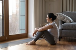 Dreamy Indian ethnicity woman in sports pants and t-shirt looks out window relax after work out training sit near heating grid with ventilation by floor in hardwood flooring. Modern home, rest concept