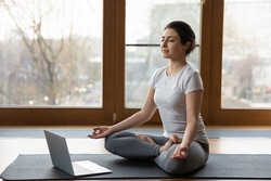 Serene Indian ethnicity woman sit cross-legged on yoga mat listen soothing music on laptop, do meditation practise with eyes closed alone indoor. Remote e coach training, modern tech, work out concept
