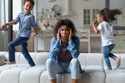 Excited naughty, noisy African children running around mom on sofa, shouting, making mother upset, annoyed. Frustrated mum sitting on couch at home, feeling headache, stress. Parenting problems