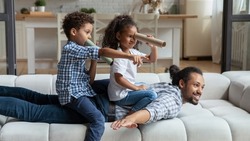 Happy African American daddy and little sibling kids playing funny active games on couch, Children sailing dad like pirate boat, holding paper toy spyglasses. Childhood, family, fatherhood