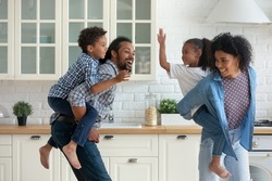 Happy active African parents piggybacking sibling children, running in kitchen, chasing each other dancing. Energetic family couple carrying kids on backs, running, playing funny games at home