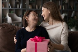 Loving young woman grown-up daughter congratulating mature mother, hugging, presenting pink gift box, sitting on couch at home, family celebrating special event birthday or mothers day concept