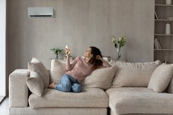 Smiling woman using air conditioner remote controller, relaxing on comfortable couch in living room alone, positive beautiful young female switching temperature on climate control system at home