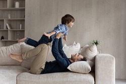 Dad lying on couch lifts on outstretched hands little 6s son. Happy cute boy play plane fly in air while his father raise him up on arms. Active weekend at home, dreams about holiday, leisure concept