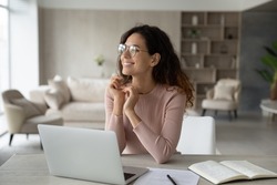 Smiling dreamy confident businesswoman in glasses looking to aside, sitting at work desk with laptop, happy pensive woman student freelancer visualizing good future, dreaming of new job opportunities