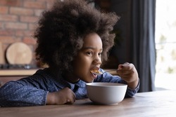 Happy hungry little adorable curly African American kid girl eating crunchy fast dry meal from bowl on breakfast, tasting yummy sweet corn flakes with milk, balanced meal for children concept.