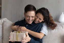Happy 30s Hispanic wife congratulates husband on birthday, woman makes surprise, gives to beloved man gift box with present, family celebrating life events, hugging expressing love. Congrats concept