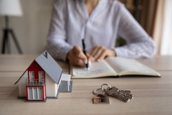 Close up young woman handwriting notes in paper organizer, sitting at table with keys and house model, household expenditures management, planning real estate investment, purchasing own accommodation.