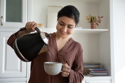 Smiling young indian ethnicity woman pouring hot water from kettle into cup, brewing tea or coffee, drinking beverage in modern kitchen, enjoying peaceful morning evening stress free carefree time.