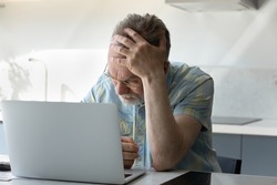 Frustrated tired senior man touching head at laptop, receiving shocking bad news, siffering from headache, migraine, memory loss, losing money due to fraud, scam, insecure financial data, hacking
